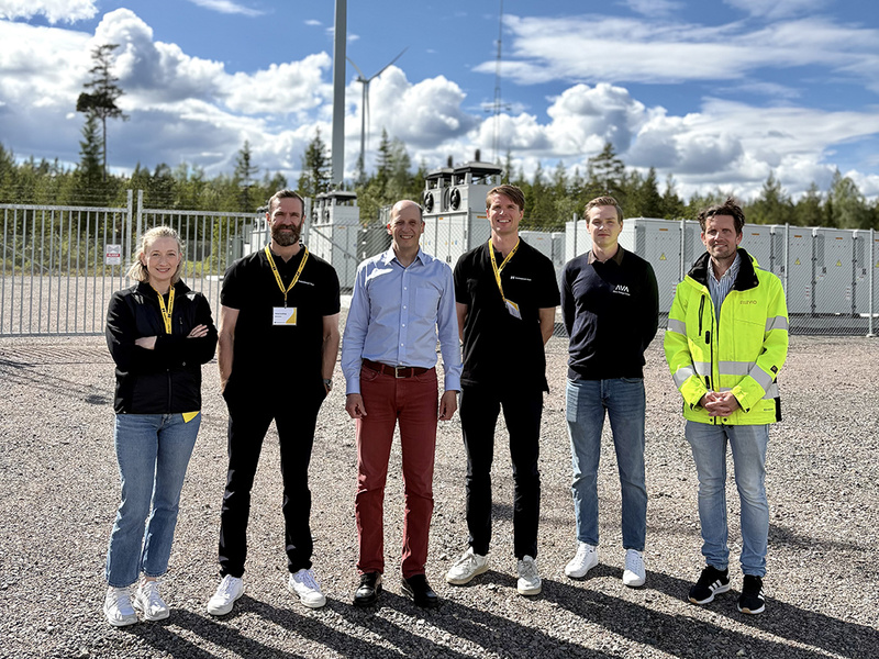En grupp människor står utomhus vid en batteri- och vindkraftpark med vindkraftverk och batterier i bakgrunden.