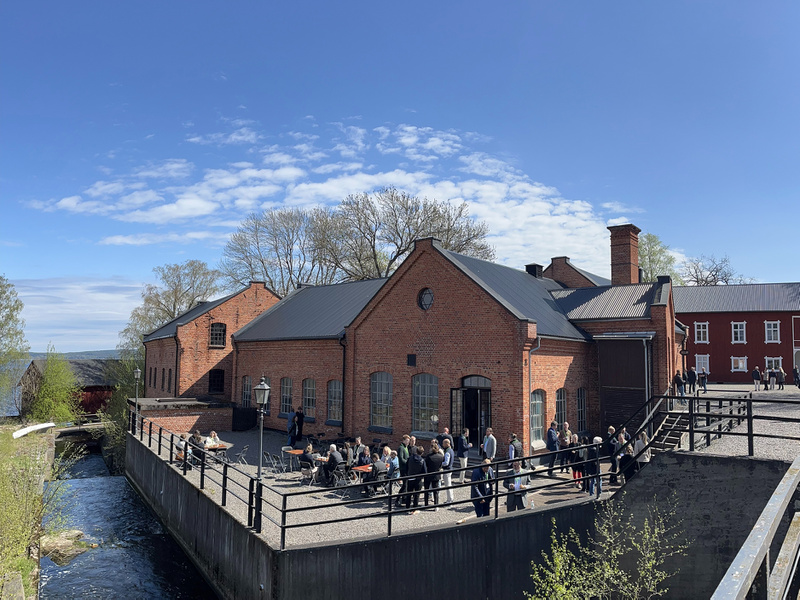 Folk minglar utanför en röd tegelbyggnad i solljuset vid en sjö.