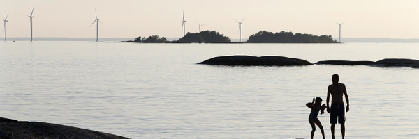 En vuxen och ett barn står på en klipphäll i vattnet vid en stilla insjö. I bakgrunden syns flera vindkraftverk ute i sjön.